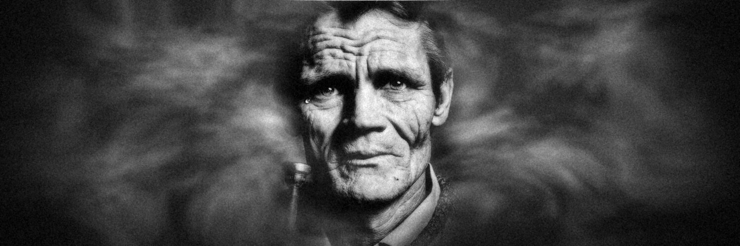 Chet Baker in 1986, close-up in zwart-wit met een traan onder zijn rechteroog en zijn trompetmondstuk tegen de wang, in rokerige mist. Foto: John Claridge, bewerkt.